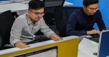 Two men collaborate on computers in an office, focusing on proposal management software for bidding processes.